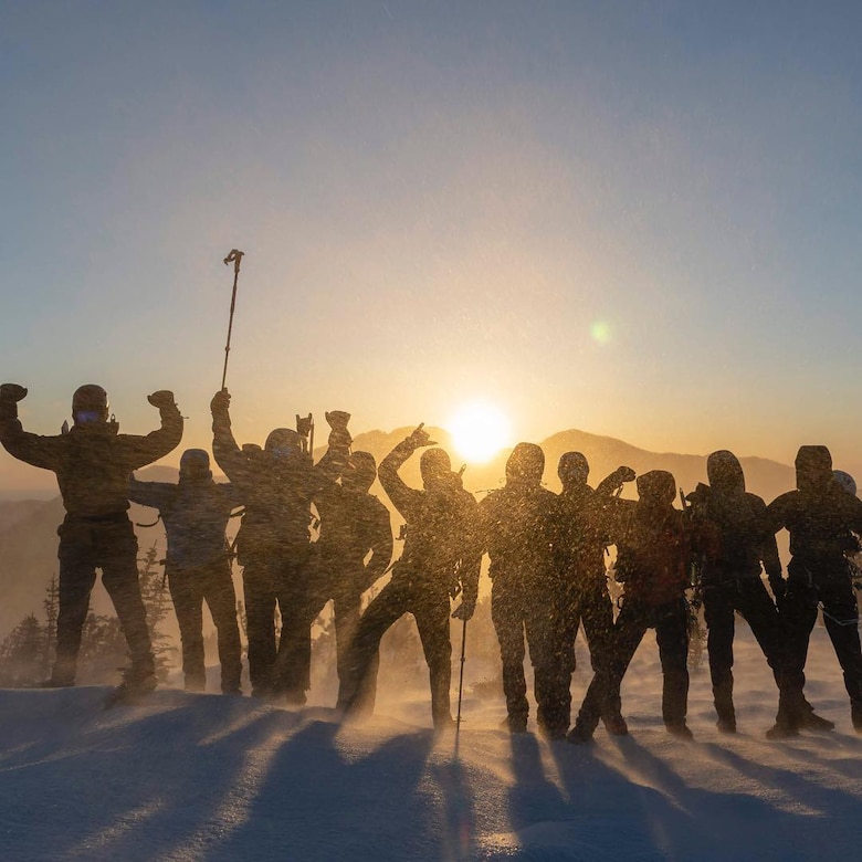 Several men standing on a mountain are backlit by the sun and more mountains.