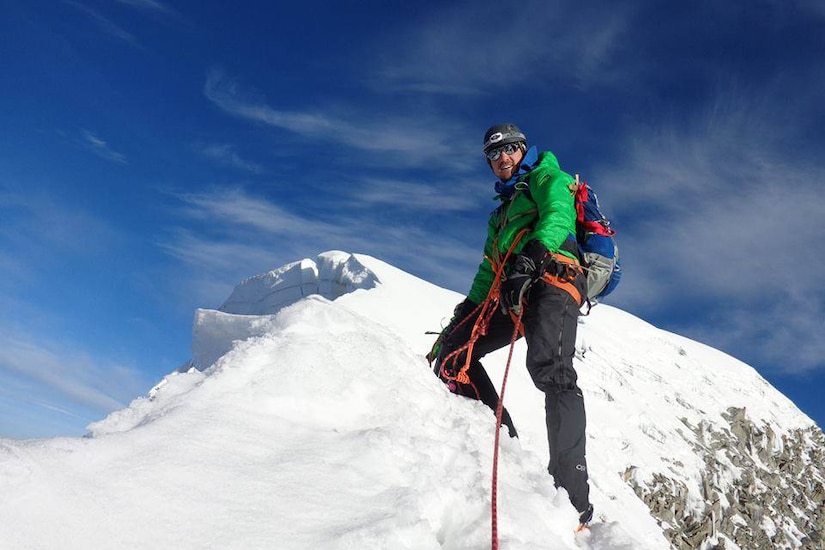 A man stands holding a tether near the top of a snowy mountain peak.
