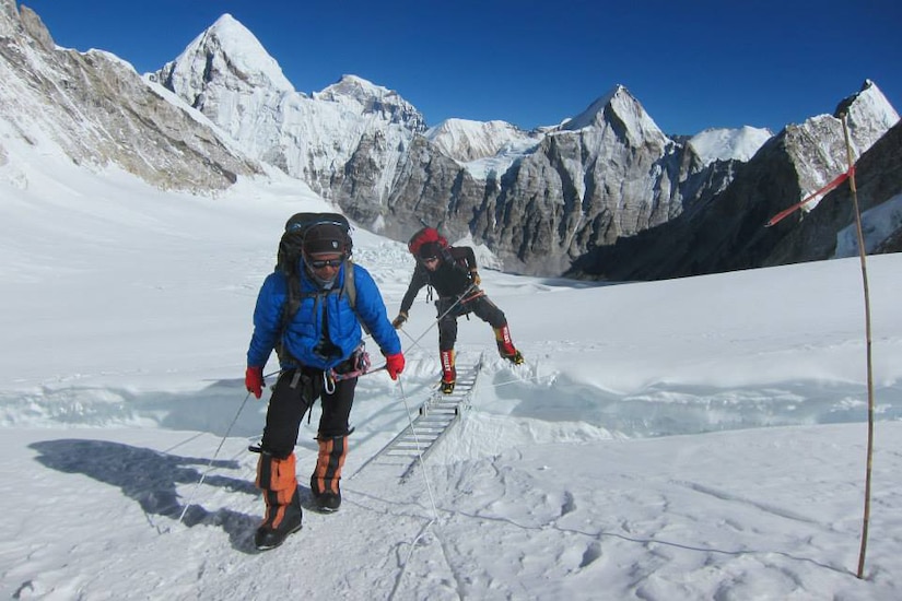 A man walks in front of a ladder bridging a snowy crevasse. A second man crosses the ladder.