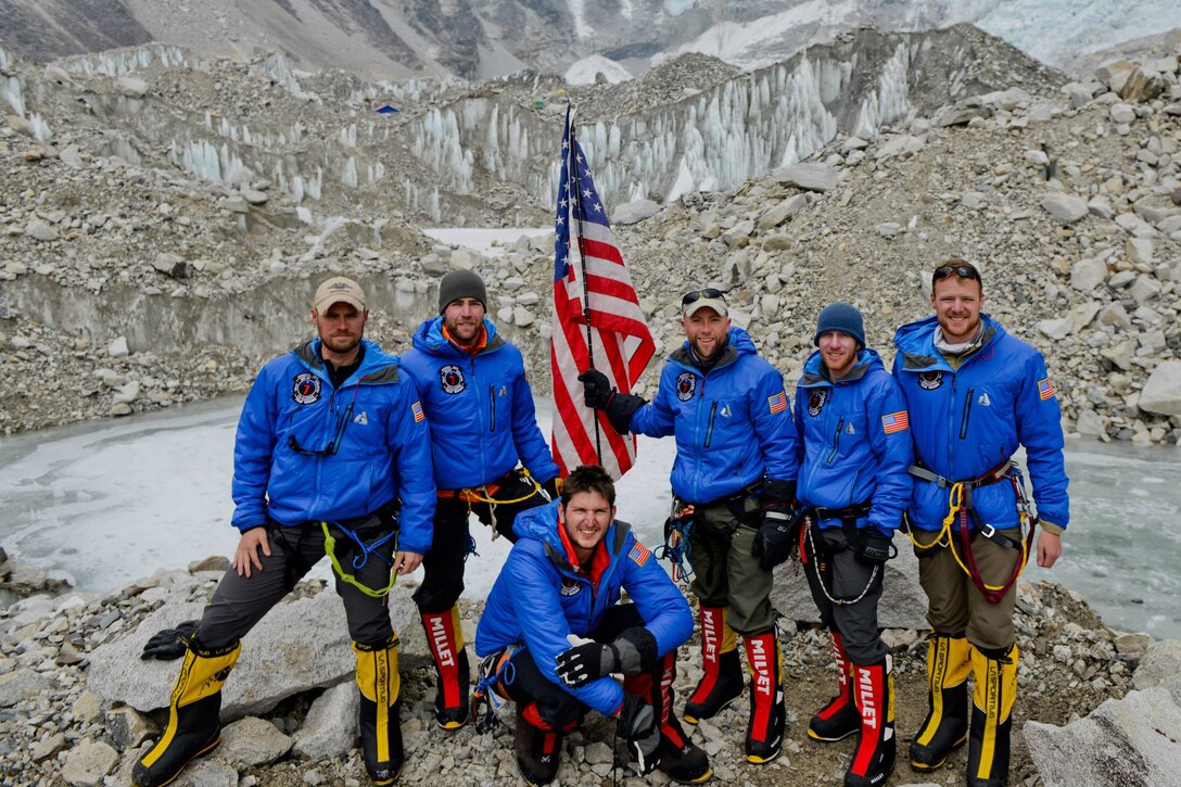 Six men pose with a U.S. flag in front of a craggy mountain.