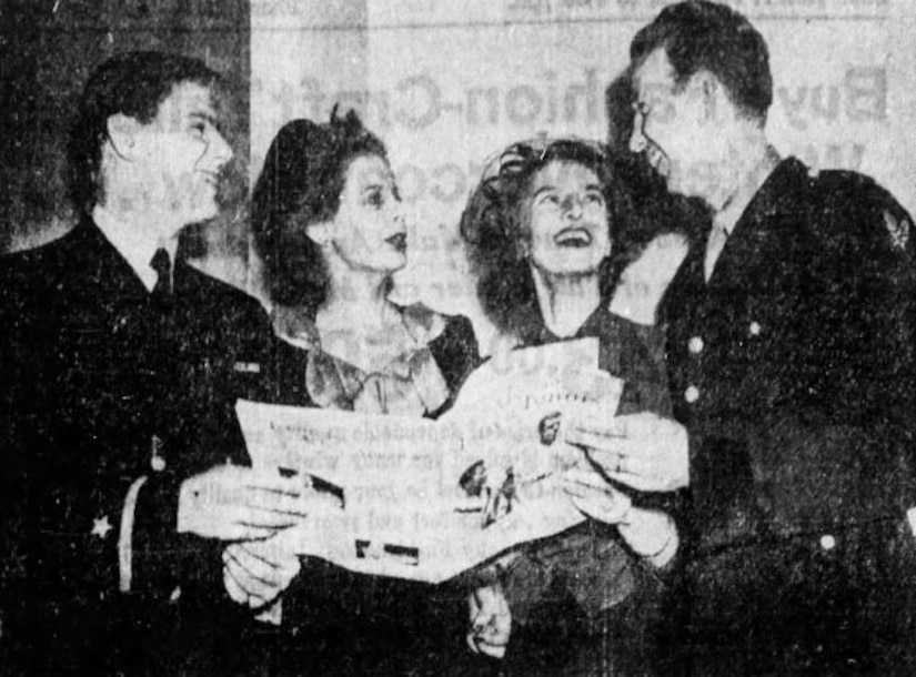 Two men wearing military uniforms smile as they hold a newspaper and talk to two women.