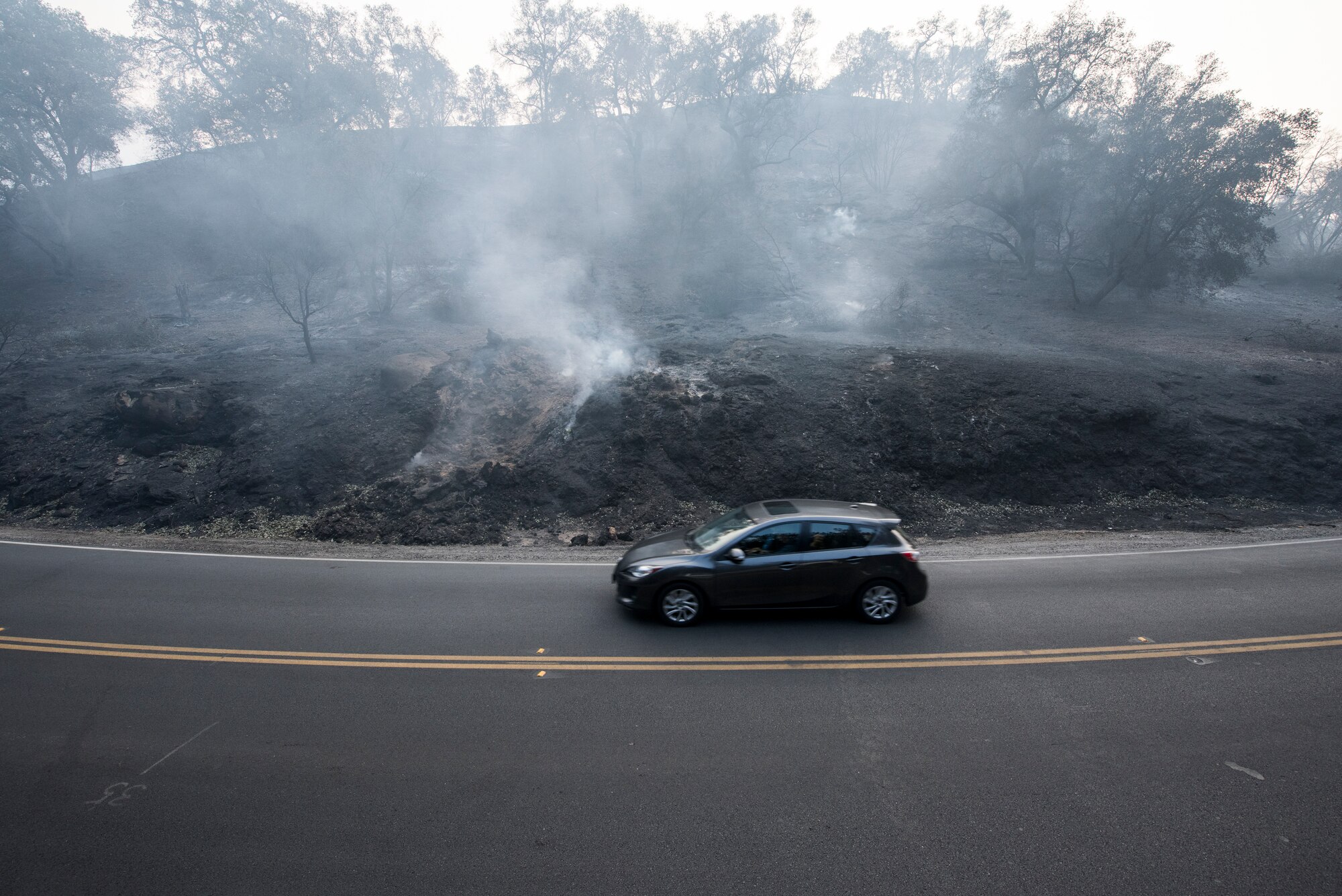 A car drives by a still burning hill in Ojai, California, Dec. 12, 2017. The Thomas Fire started on Dec. 4, 2017 in Santa Paula, near Thomas Aquinas College. Driven by Santa Ana winds gusting up to 70 mph, the flames screamed across the hillsides toward Ojai, Ventura and Santa Barbara. The 146 Airlift Wing was activated Dec.5, 2017, to support CAL FIRE with wildfire suppression efforts within the state. (U.S. Air Force photo/Master Sgt. Brian Ferguson) (U.S. Air Force photo/Master Sgt. Brian Ferguson)