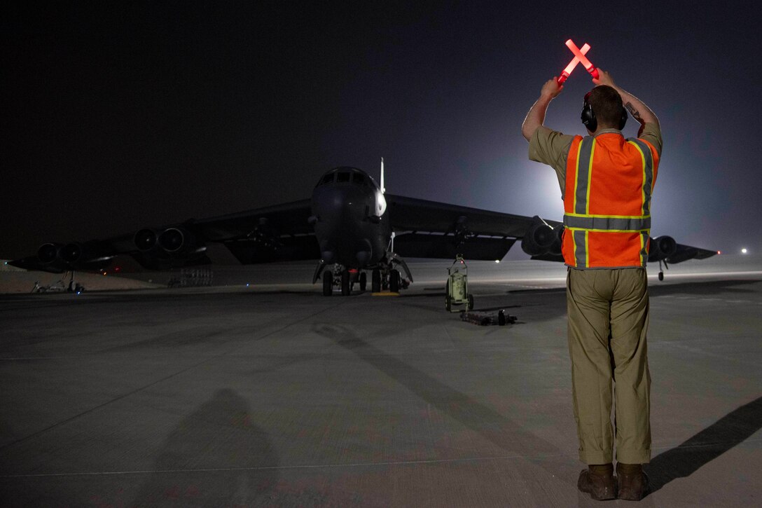 A military aircraft taxis on an airfield.  A man uses a light to direct the aircraft.