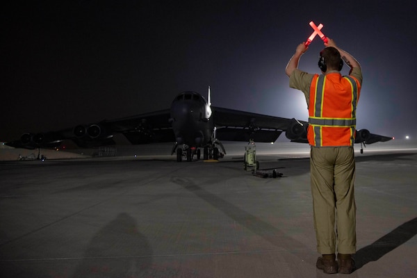 A military aircraft taxis on an airfield.  A man uses a light to direct the aircraft.