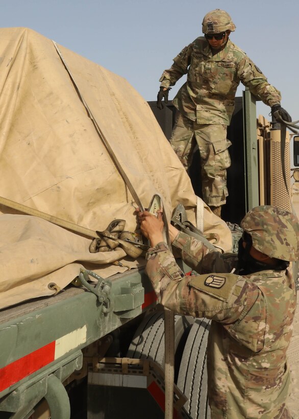 A man in a military uniform adjusts straps that are wrapped around a tarp-covered load on a truck.  Another man in a military uniform stands on the bed of the truck.