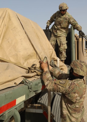 A man in a military uniform adjusts straps that are wrapped around a tarp-covered load on a truck.  Another man in a military uniform stands on the bed of the truck.