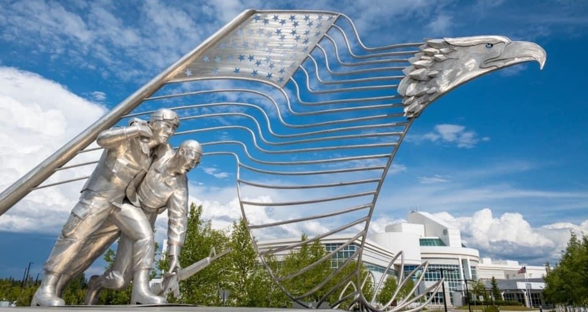 Flag made of airplane metal with eagle head as a backdrop to two soldiers. Blue sky and white hospital in background.