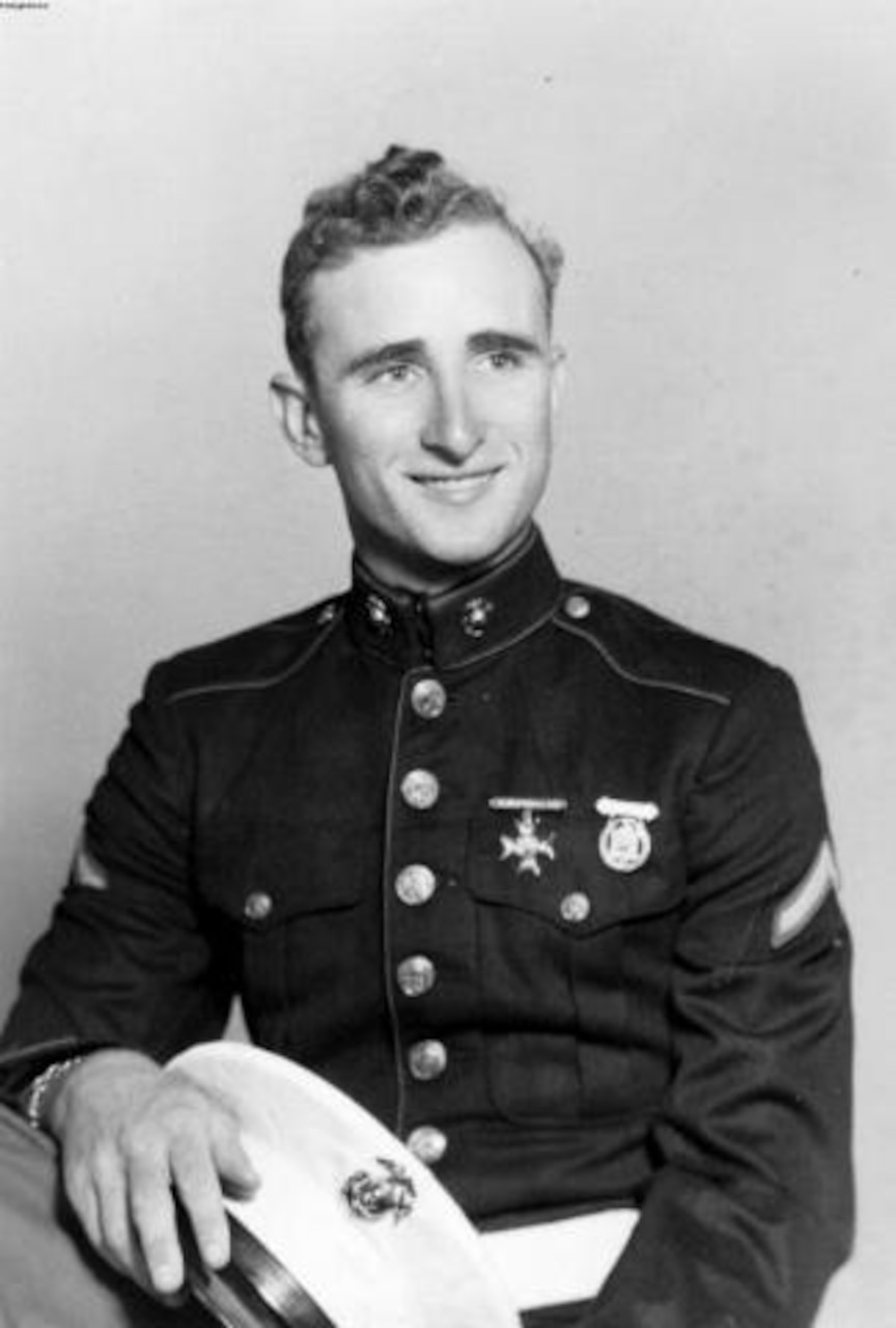 A young man in uniform smiles while holding a dress cap in his hands.