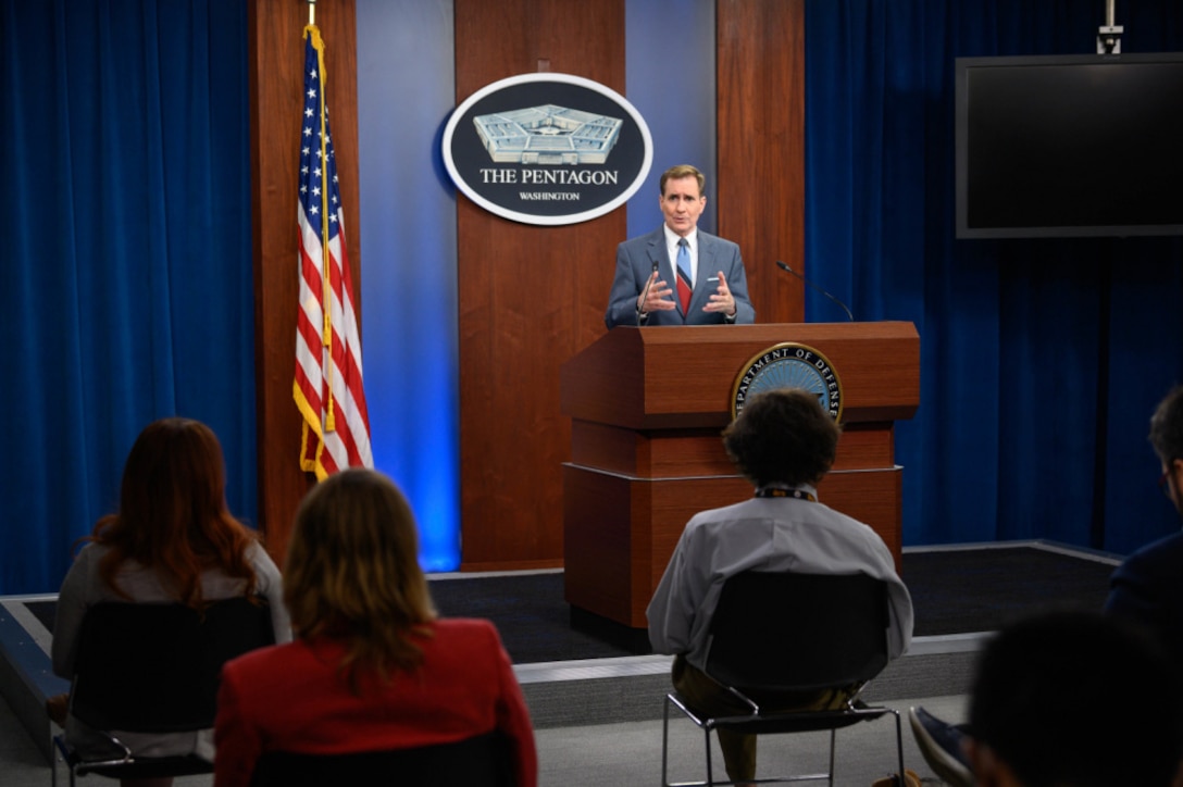 A man stands at a lectern while briefing seated  reporters.