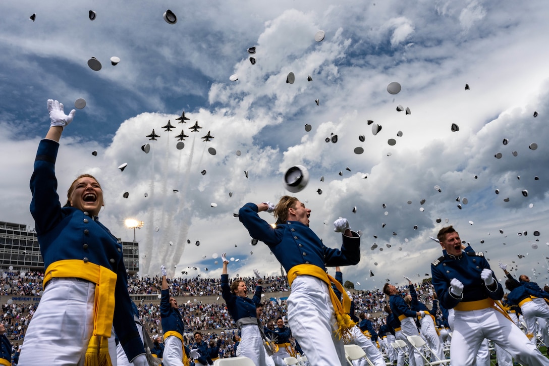 Students in military uniforms throw their caps into the air. Overhead, military aircraft fly through the air.