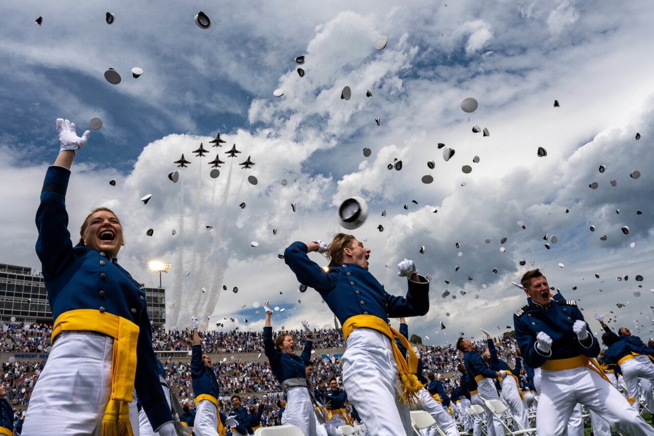 Students in military uniforms throw their caps into the air. Overhead, military aircraft fly through the air.
