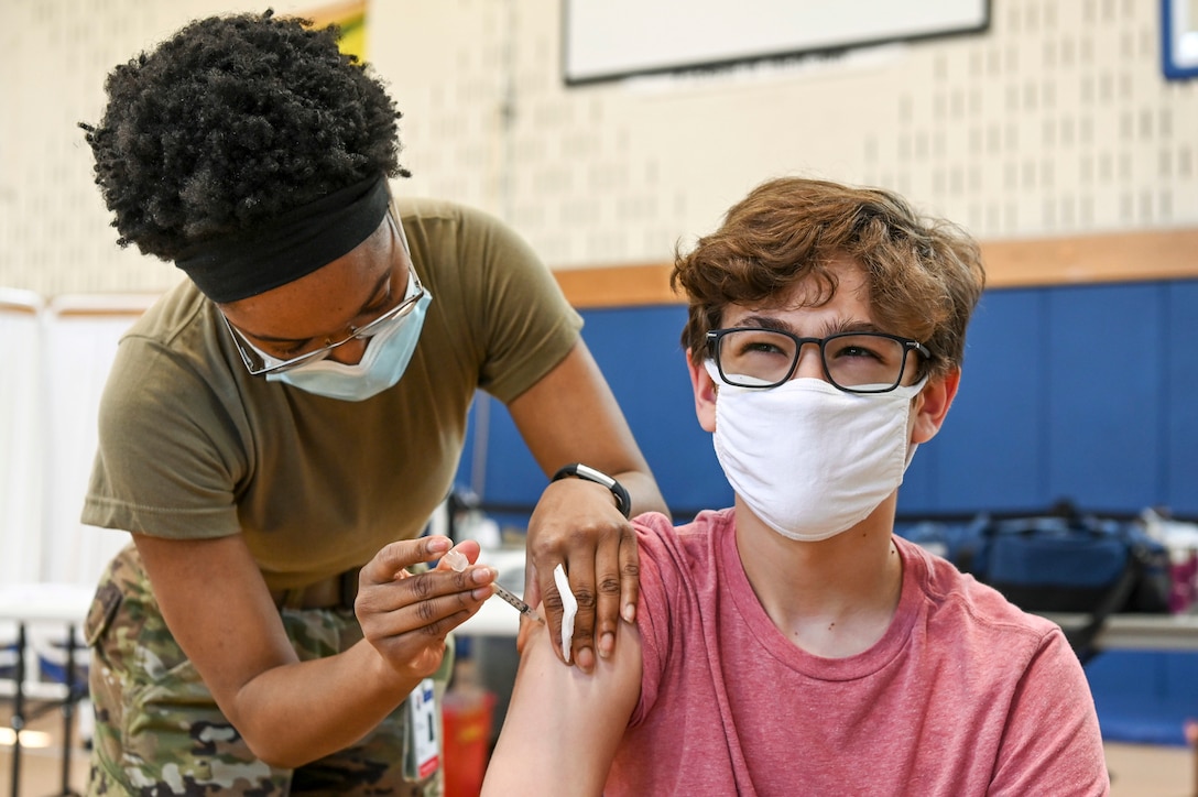 A 13-year-old wearing a mask smiles while receiving a shot from an airman.