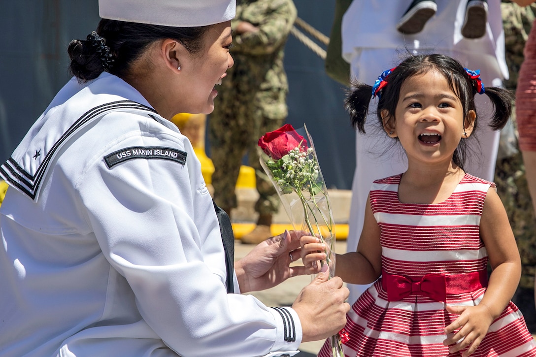 A smiling sailor kneels next to a smiling child as they both hold onto the same small flower bouquet.