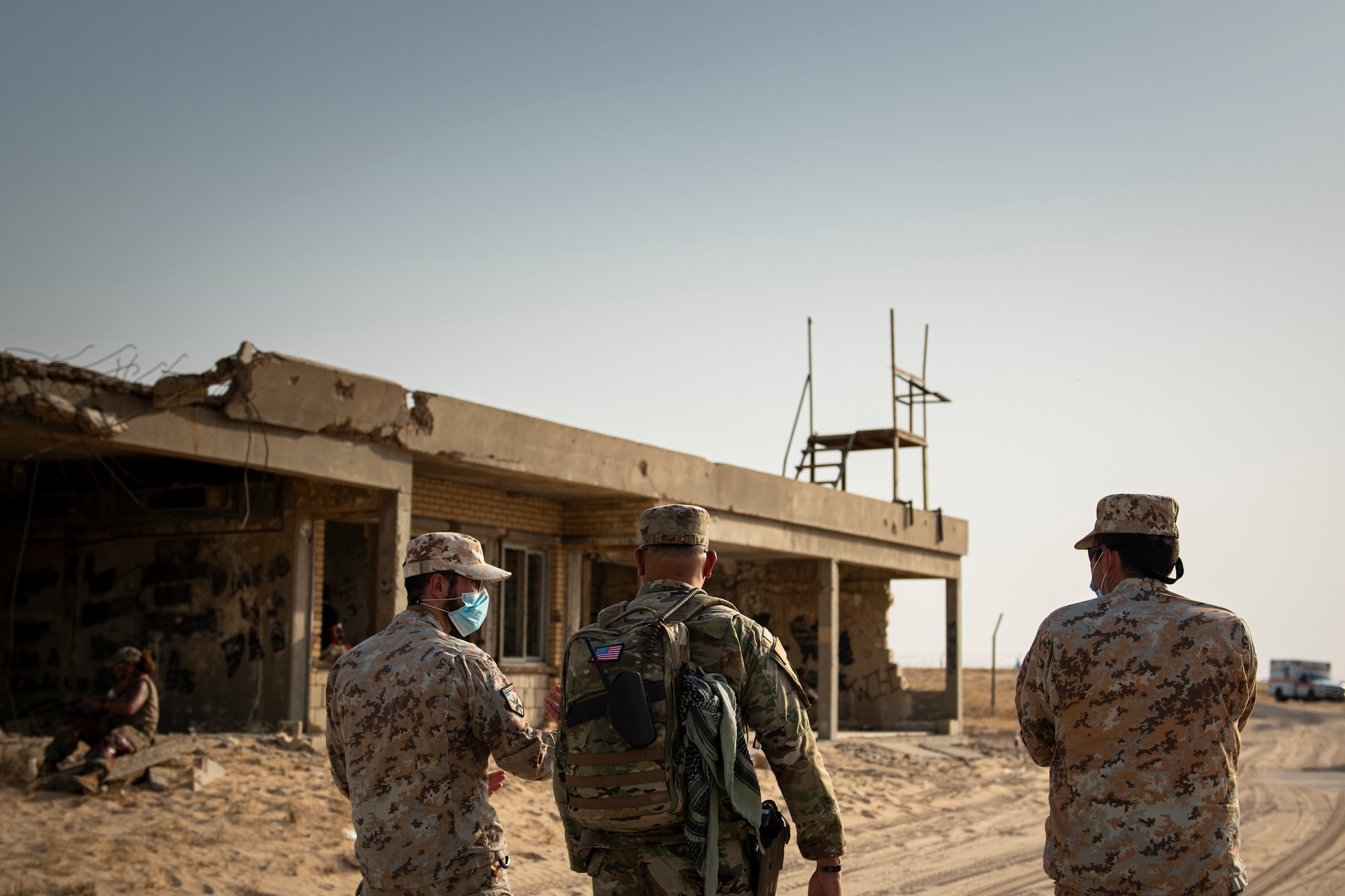 People walk along the desert next to deserting buildings.