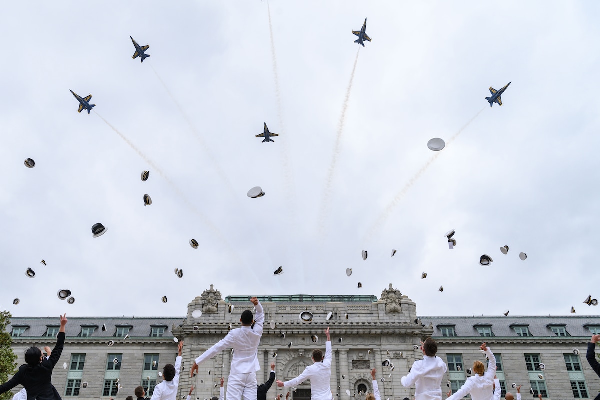 Midshipmen throw their hats in the air as five military jets fly above them.