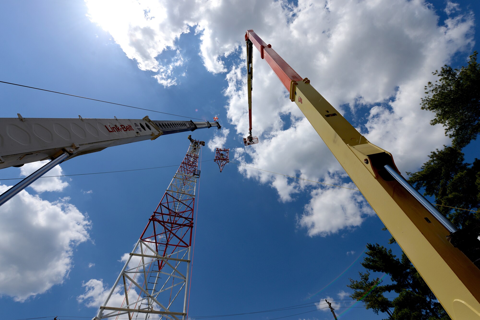 An image of Airmen with the Massachusetts and Pennsylvania Air National Guard working in unison as they use climbing gear to secure themselves safely to a tower, operate a high lift for inspection purposes and use a mobile crane to hoist and connect the top section of a radio communications tower.