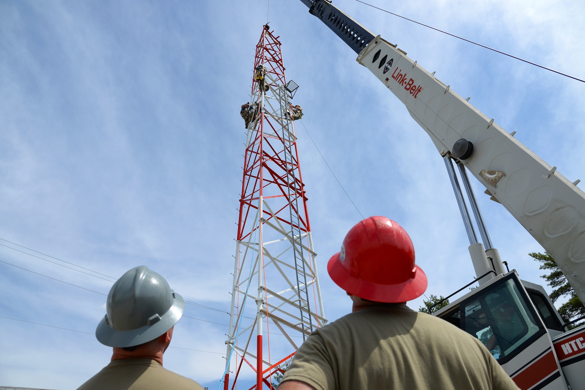 An image of Airmen with the Massachusetts and Pennsylvania Air National Guard using climbing gear and operating a mobile crane to hoist and connect a section of radio communications tower.