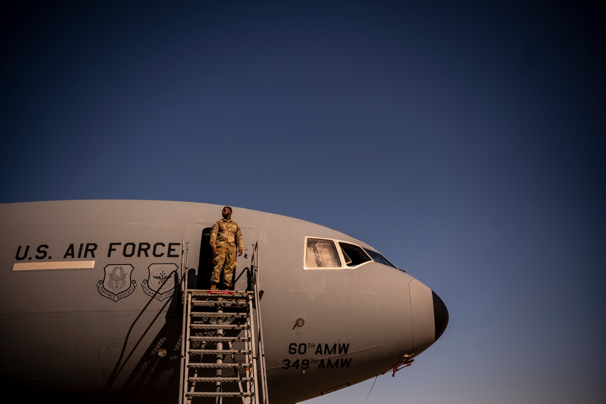 A men stands outside a large military aircraft on the top of a set of metal stairs.