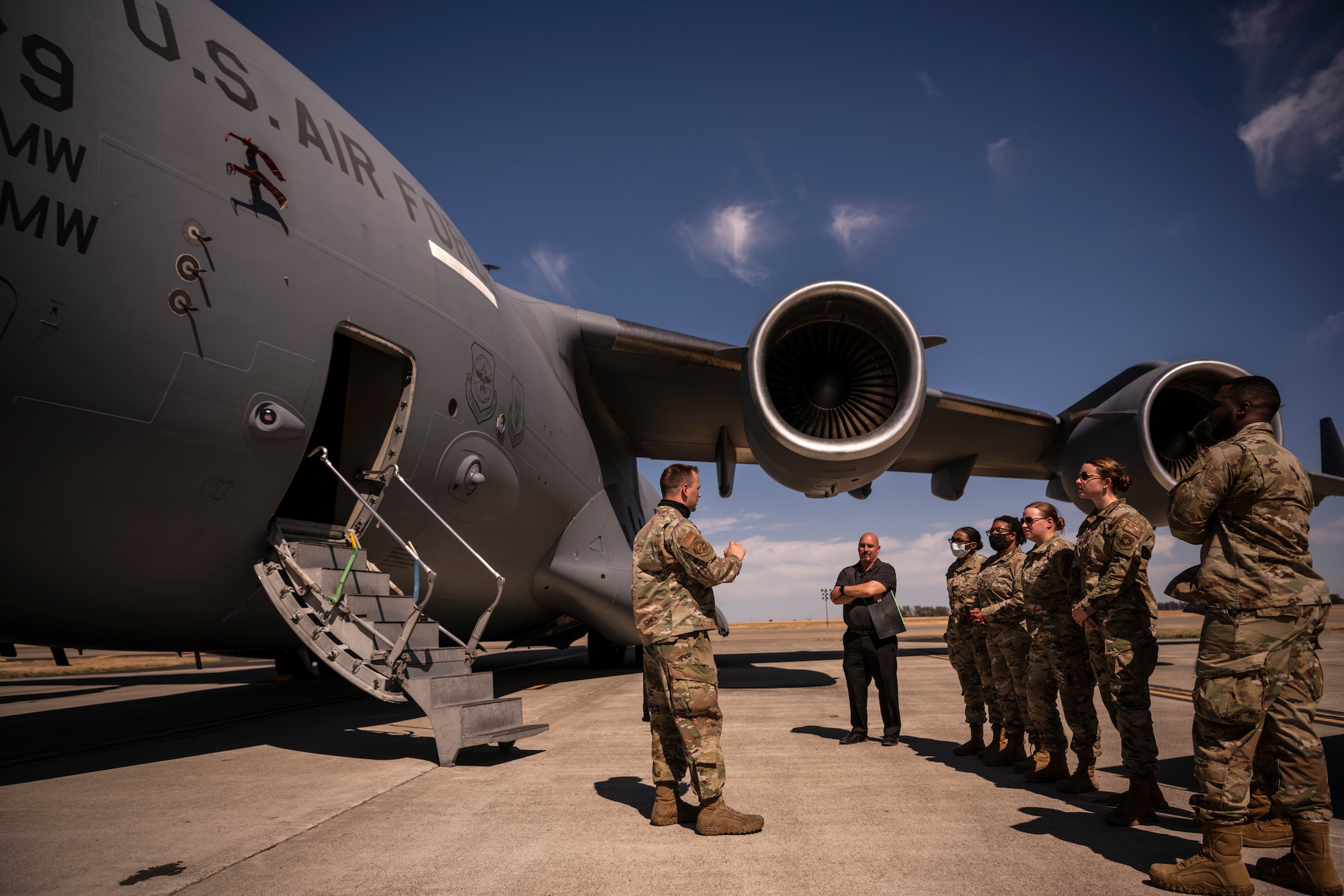 A group of Airmen stand outside of a large military aircraft on the flight line.