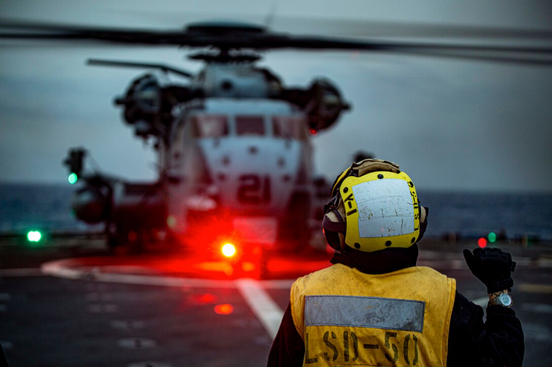 A sailor signals to a helicopter parked on a ship at sea illuminated by colorful lights.