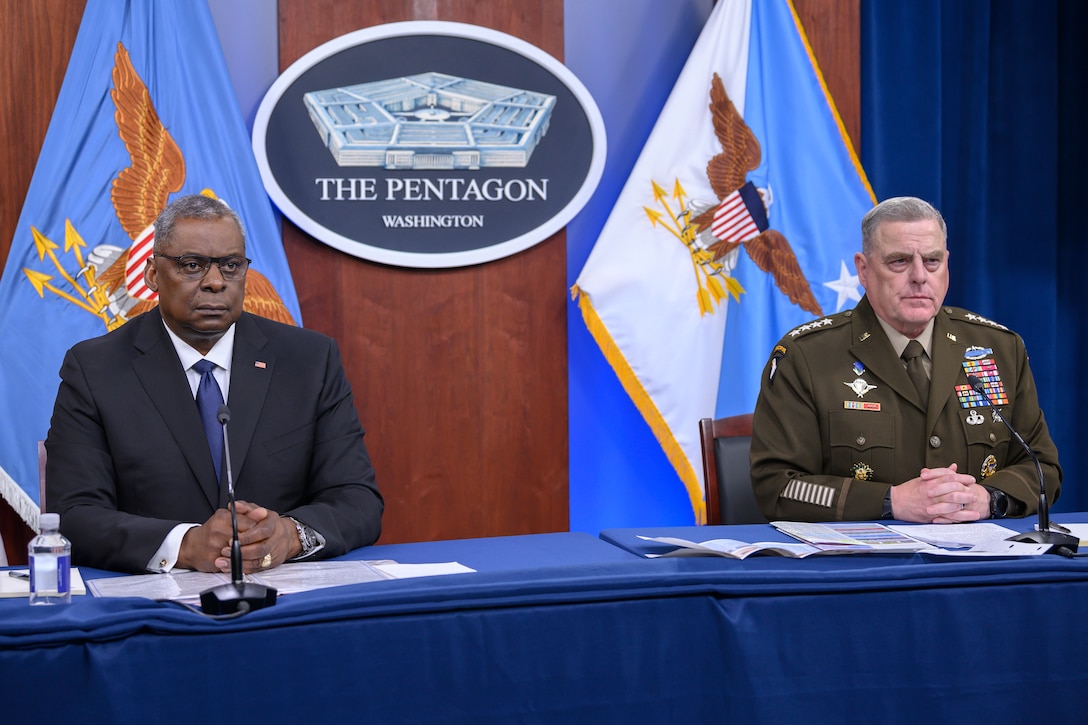 Two men sit behind a table with their hands folded in front of them.