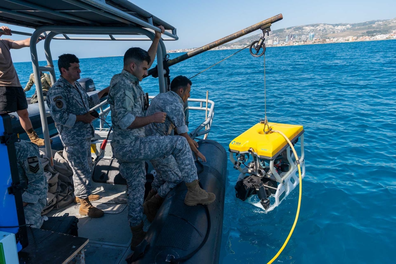 Several uniformed personnel stand at the edge of a small watercraft and operate a crane, which is being used to remove a small submersible vehicle from the water.