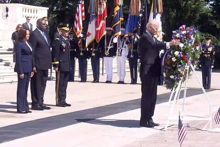 A man places his hand on a wreath, which is hanging on a metal stand.  Behind him, three people stand side-by-side with their hands at their side.