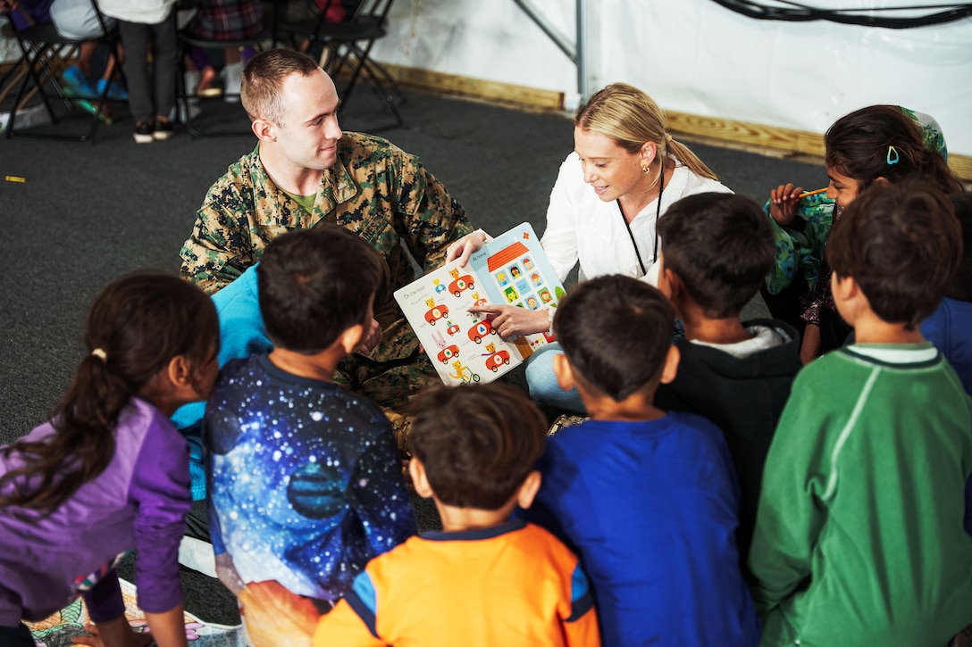 A woman points at the page of a book as a Marine and a group of children watch.