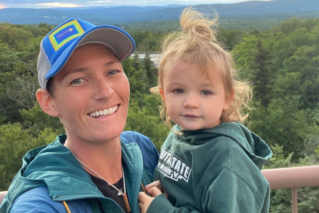 A woman smiles for a photo while holding a toddler, with mountains in the background.