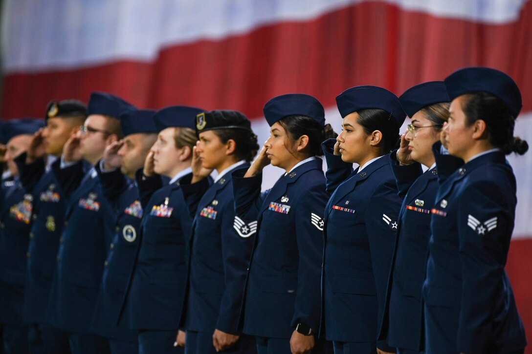 Airmen salute while standing in a straight line in front of an American flag.