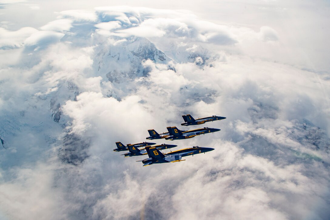 Several aircraft fly in formation over cloud-covered, snow-capped mountains.