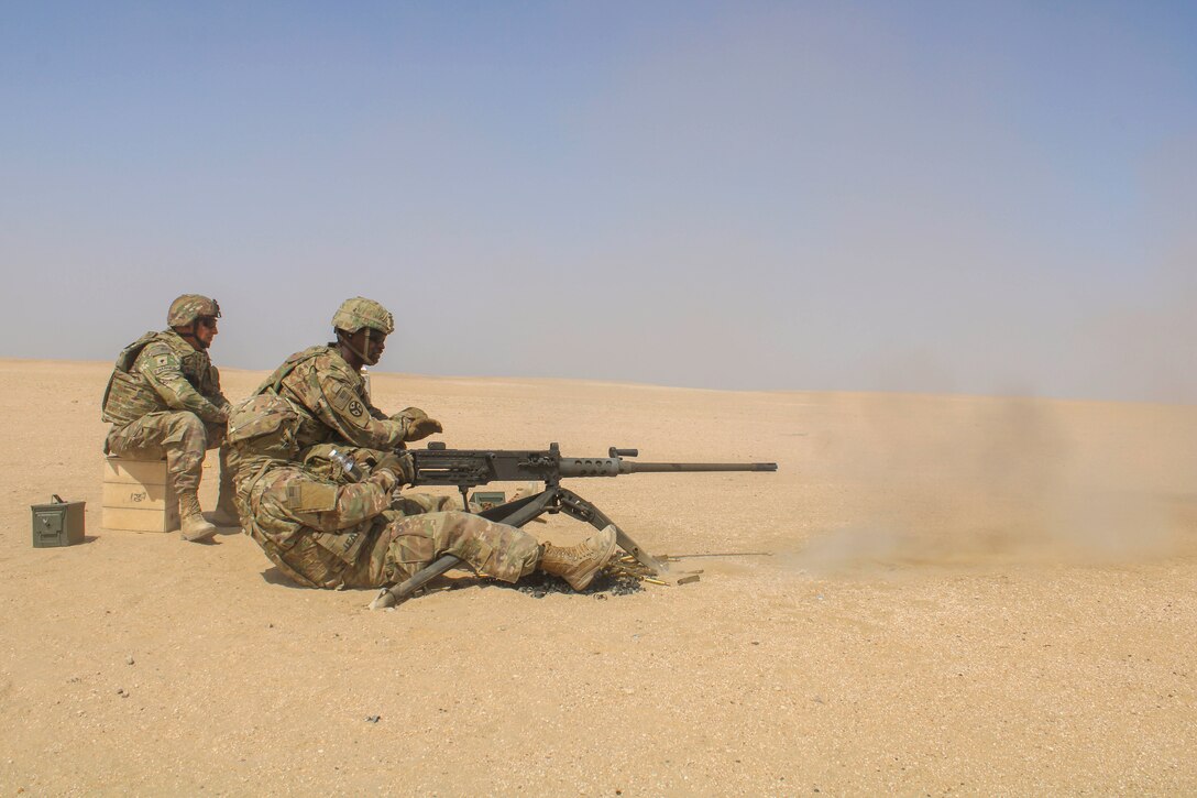A soldier leans back while firing a weapon as two fellow soldier watch in the desert.