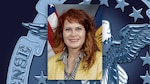 White woman with red hair and yellow jacket poses for a head/shoulders photo in front of the US flag.