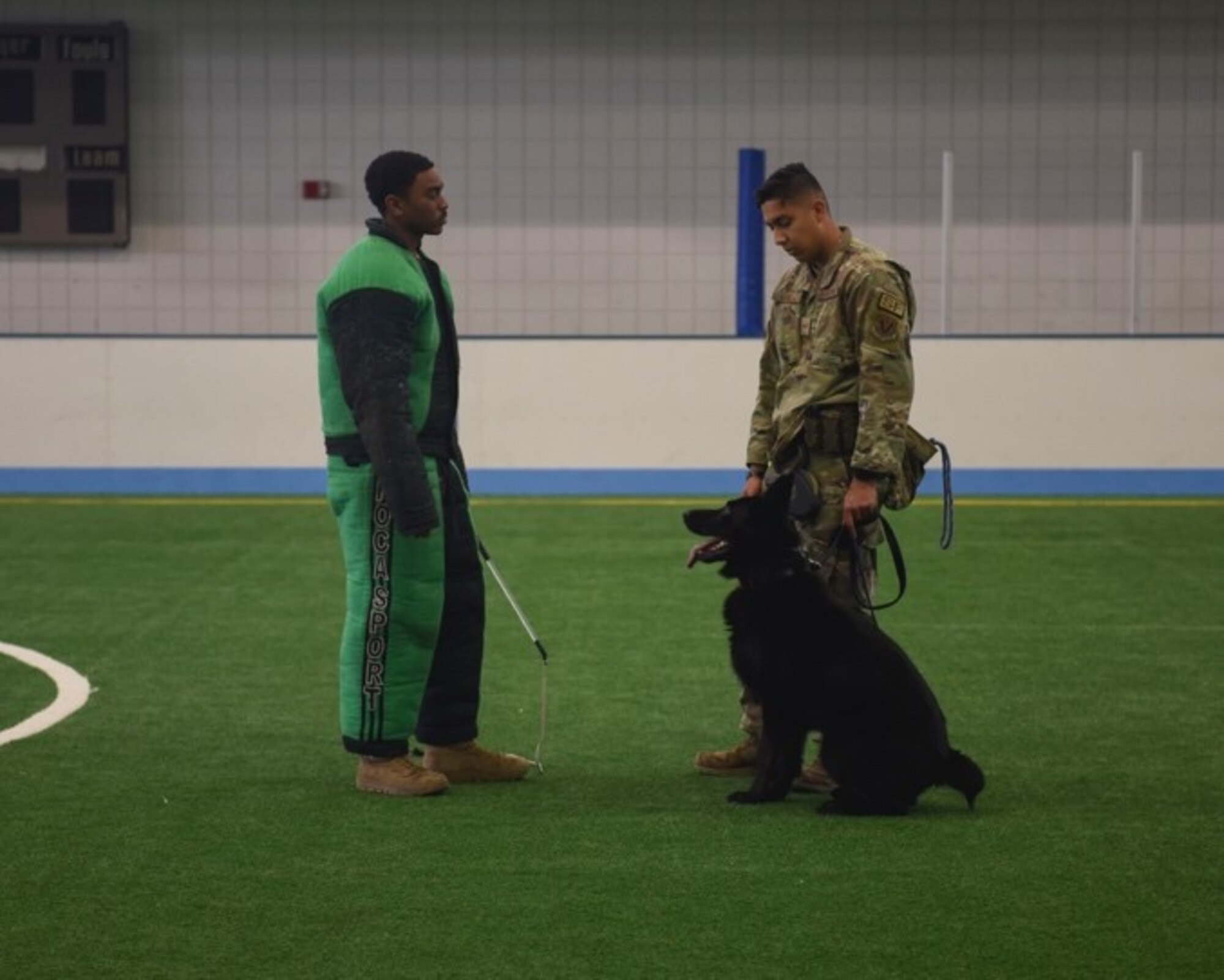 Two Airmen stand facing each other with a Military Working dog sitting at the feet of the one standing on the right as they prepare for a demonstration.