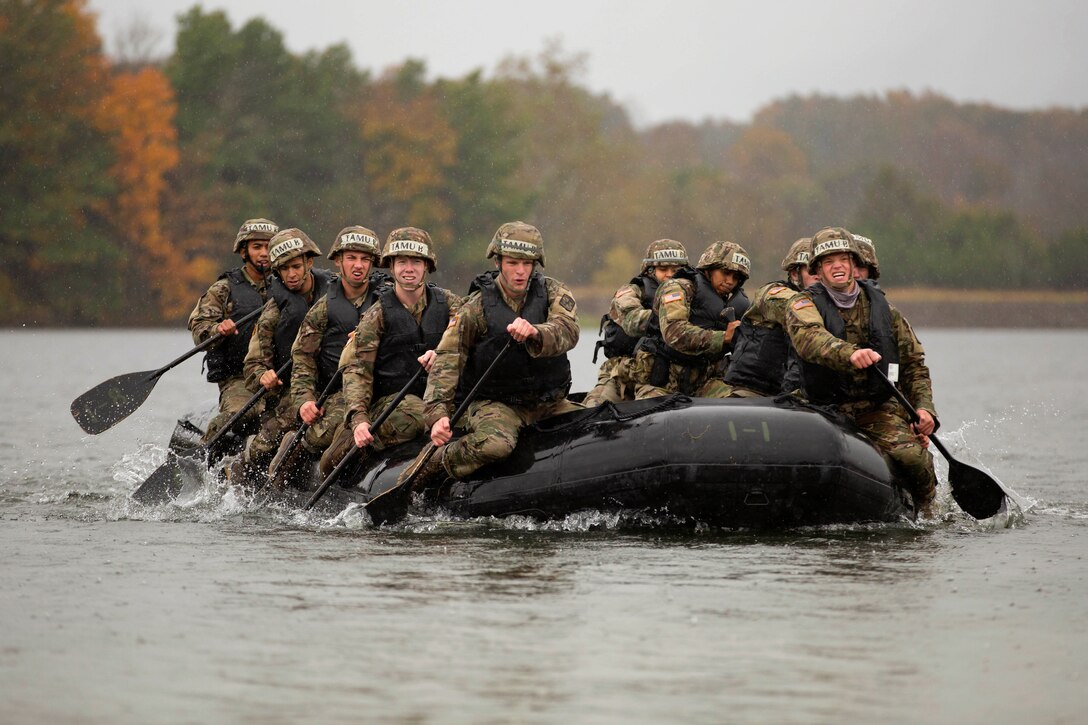 Army cadets use oars to move a watercraft in a lake surrounded by colorful trees.