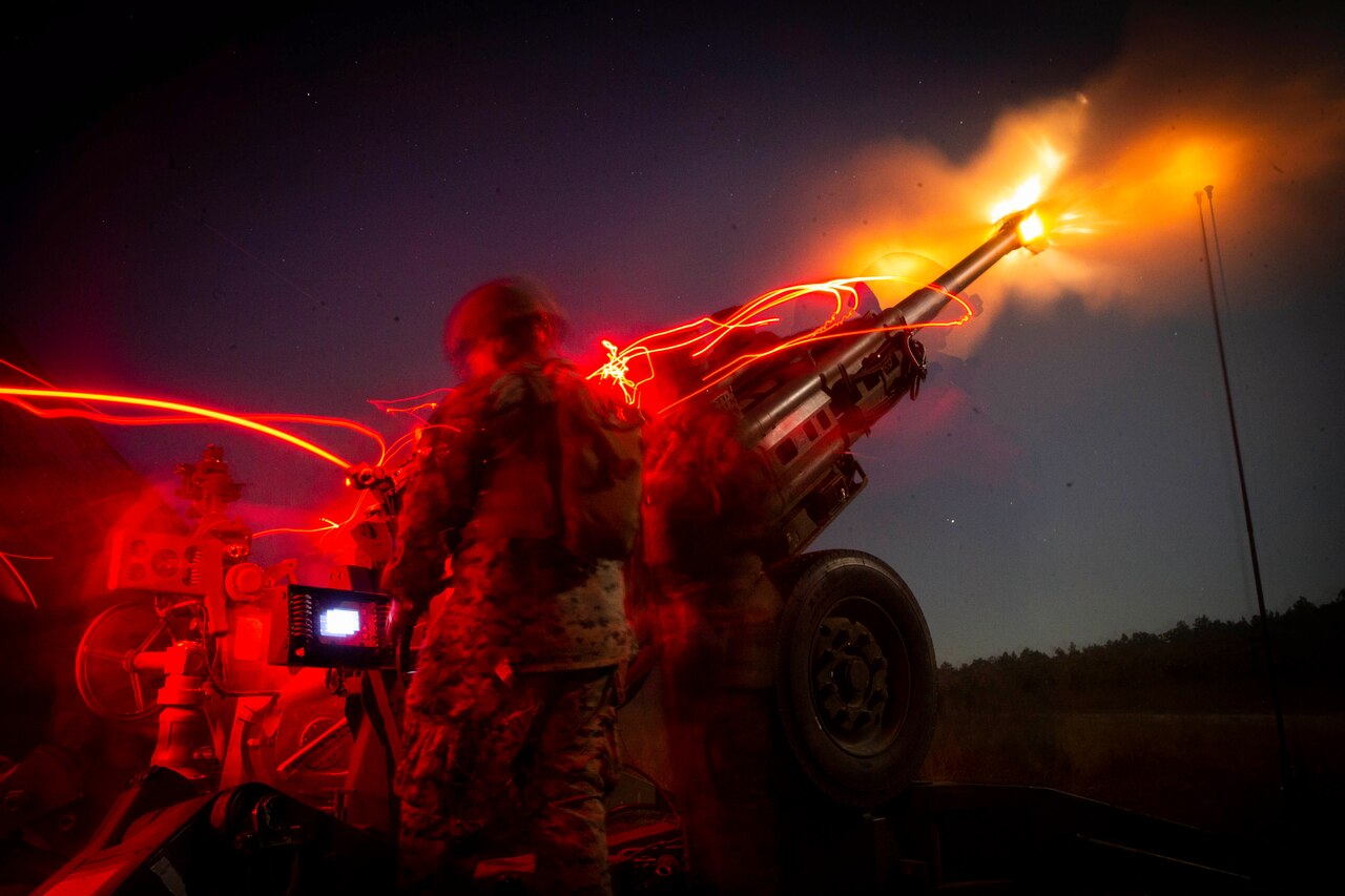 Marines fire a howitzer in the woods illuminated by colorful lights.