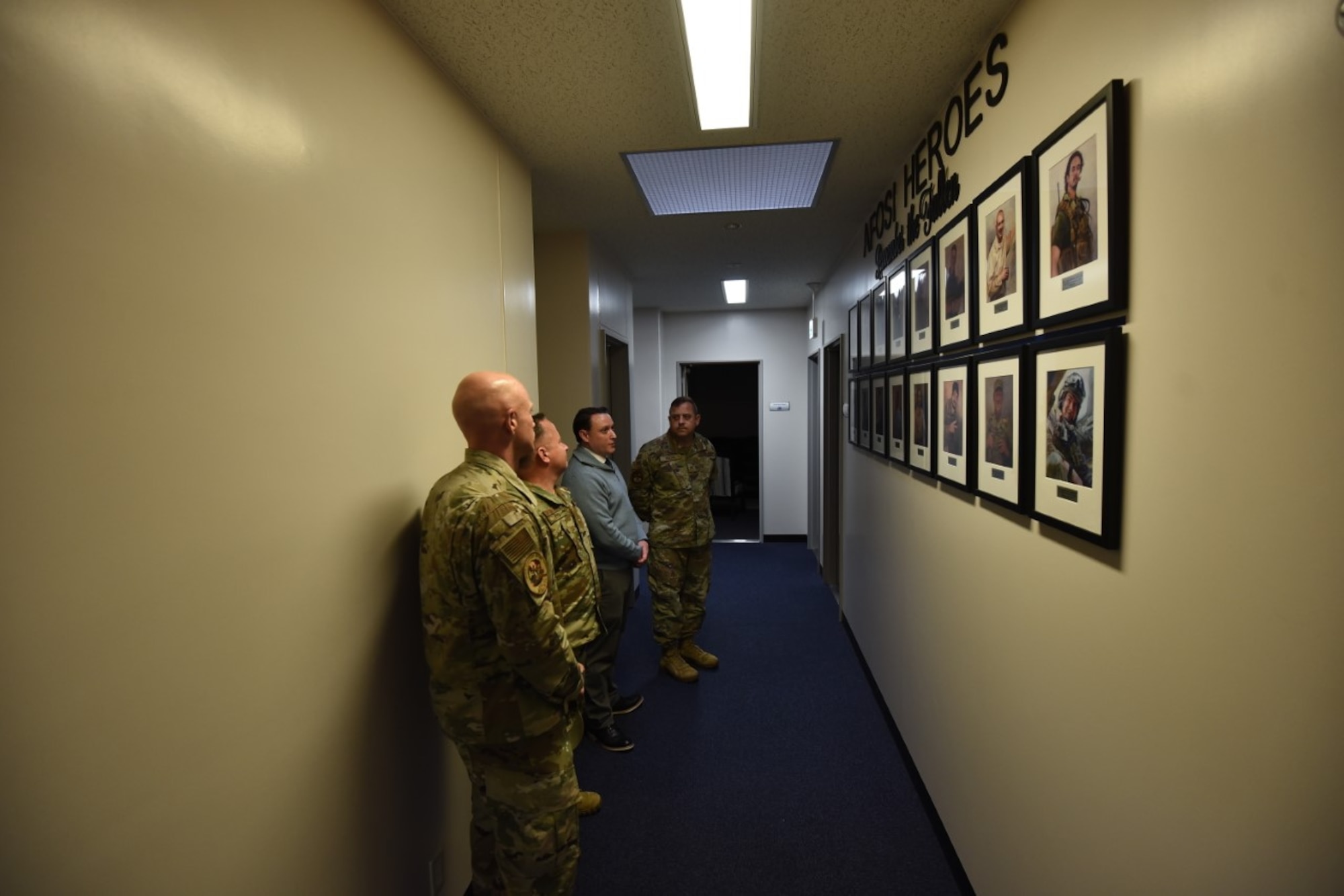 Military members stand together in front of a wall of pictures in frames of people.