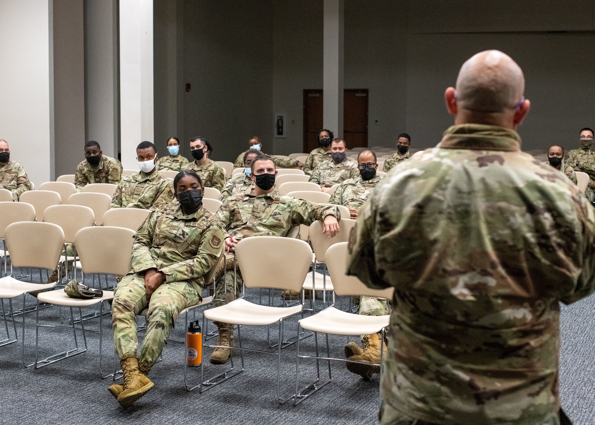 Approximately 15 Airmen sit in a room, as an Airman stands in the foreground speaking to them.