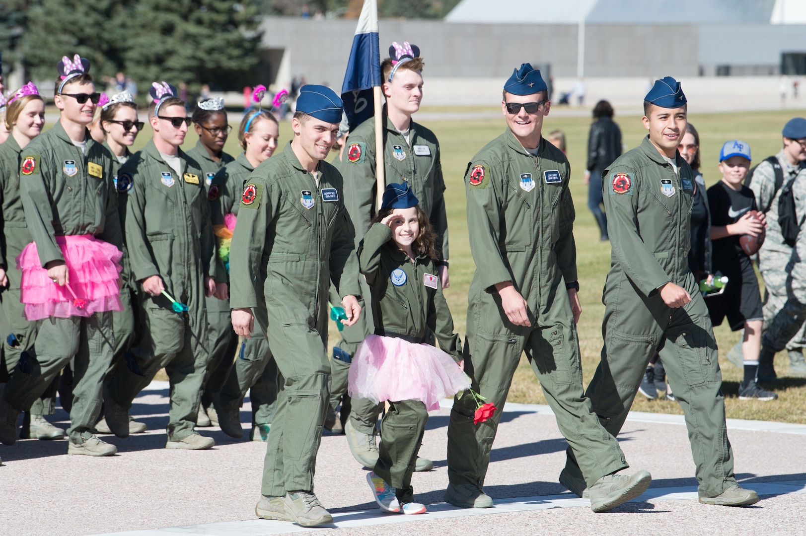 A little girl in a flight suit and pink tutu marches in an outdoor formation with cadets also in flight suits.