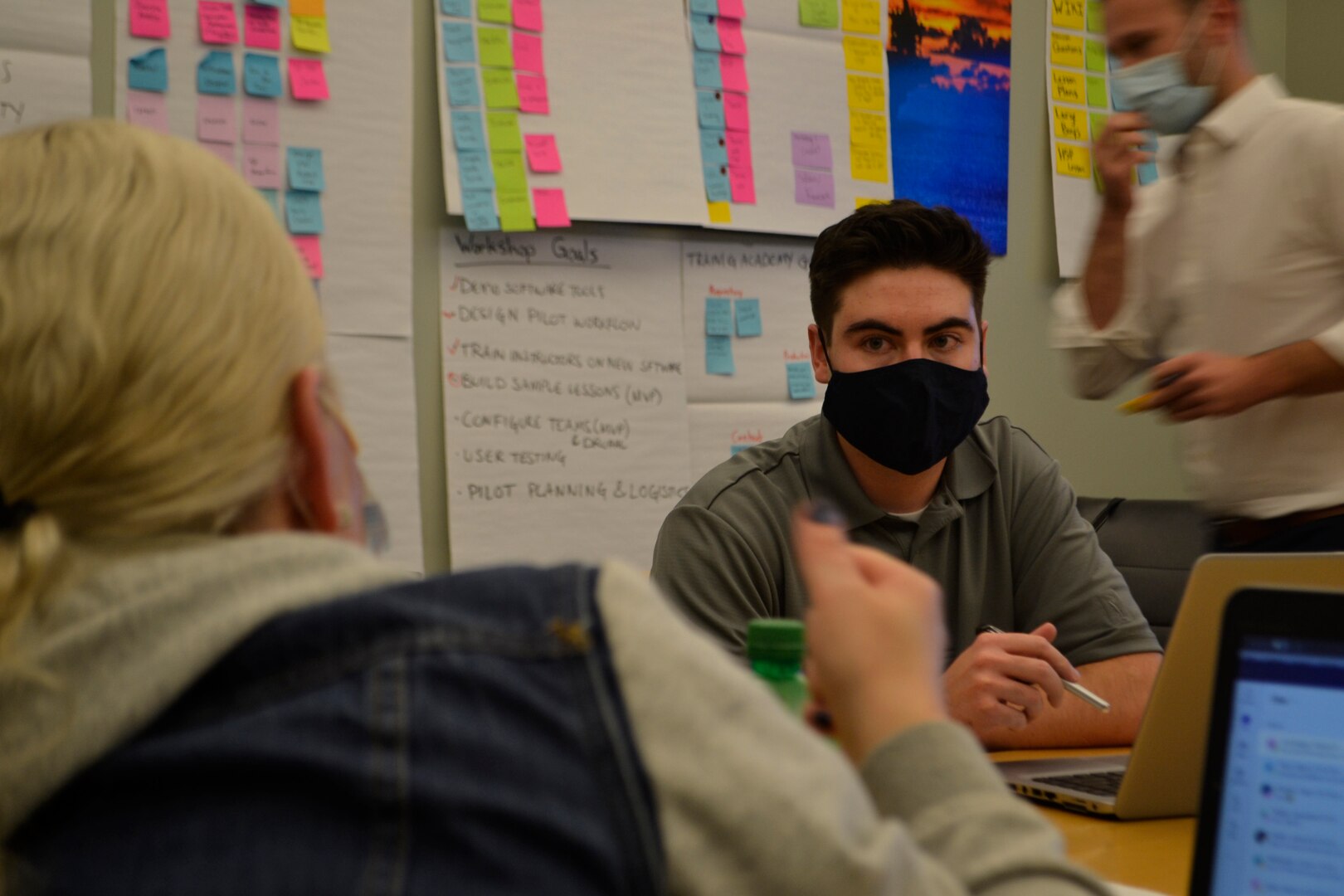 A man, right, listens as a woman, left foreground, speaks. The wall in the background is covered in colored post-it notes.