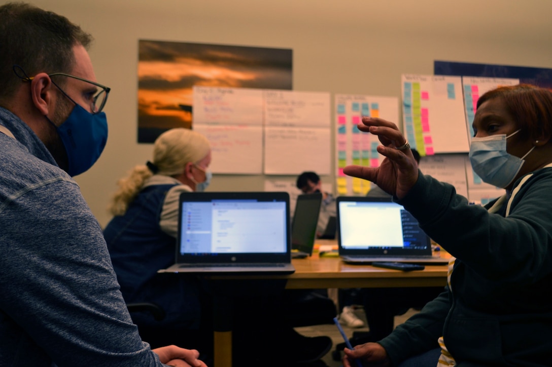 A man, left, listens as a woman, right, speaks while gesturing. There are computer screens and a wall covered in post-it notes in the background.