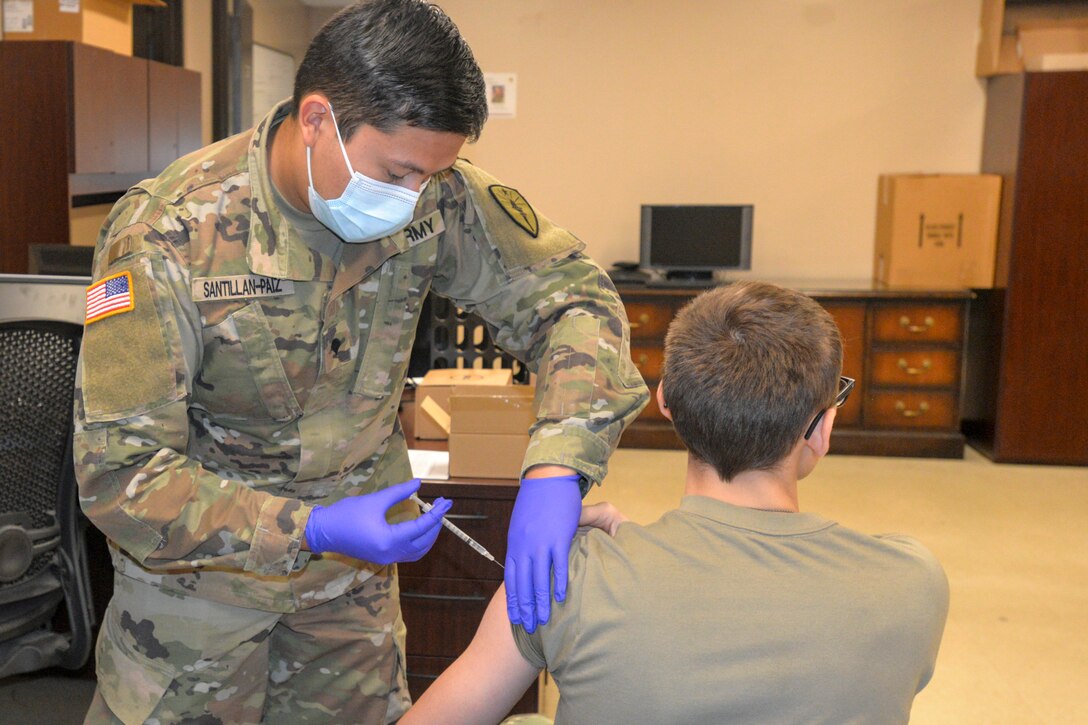 A soldier wearing a face mask and disposable gloves gives a shot to another soldier.