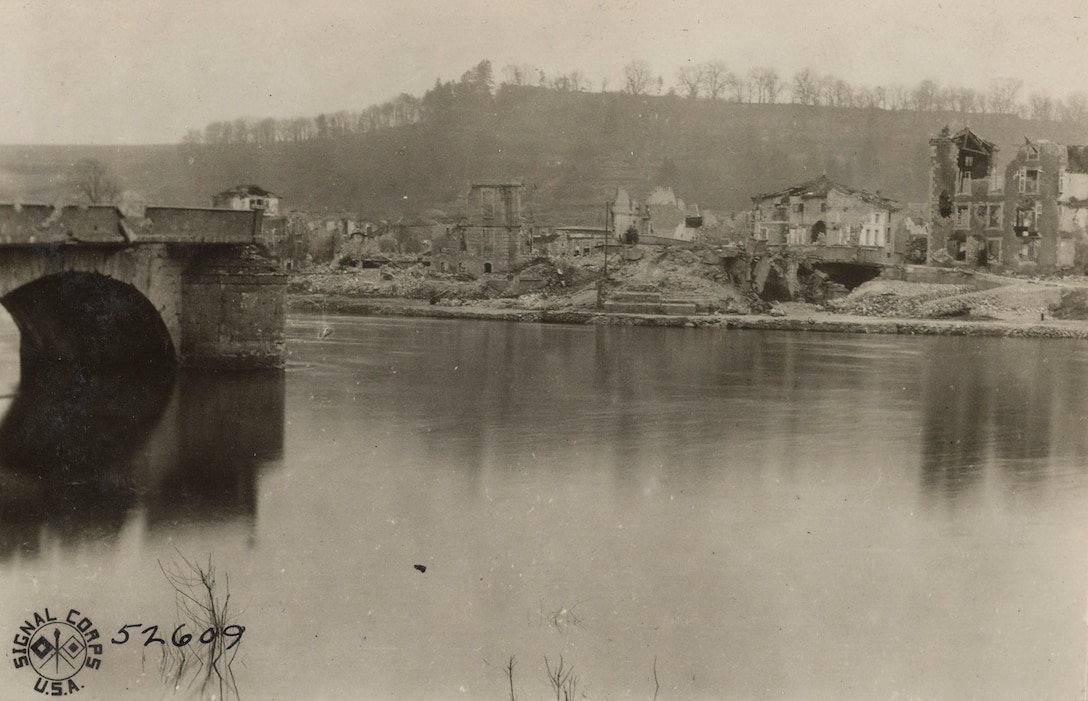 Destroyed buildings sit on the bank of a river. A destroyed bridge that doesn't reach that bank is also in view.