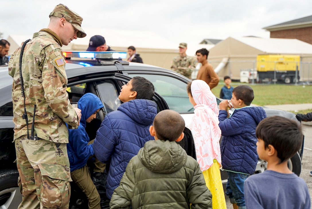 A soldier stands by a police car and talks to a group of children.