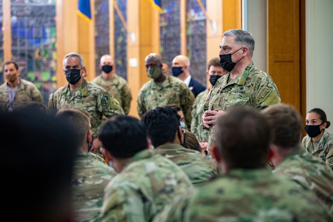 A man dressed in a military uniform and wearing a face mask stands facing a group of service members.