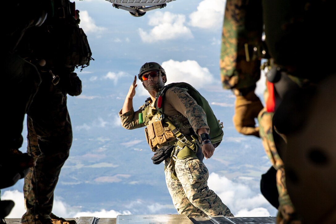 A Marine salutes as he jumps out of an aircraft wearing a parachute.