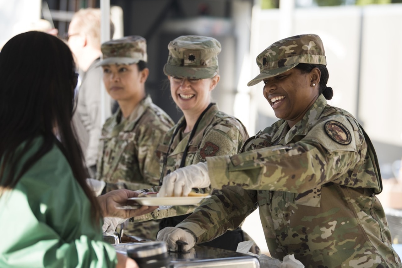 A woman in military uniform serves another woman food. Two women in uniform next to her wait to serve food as well.