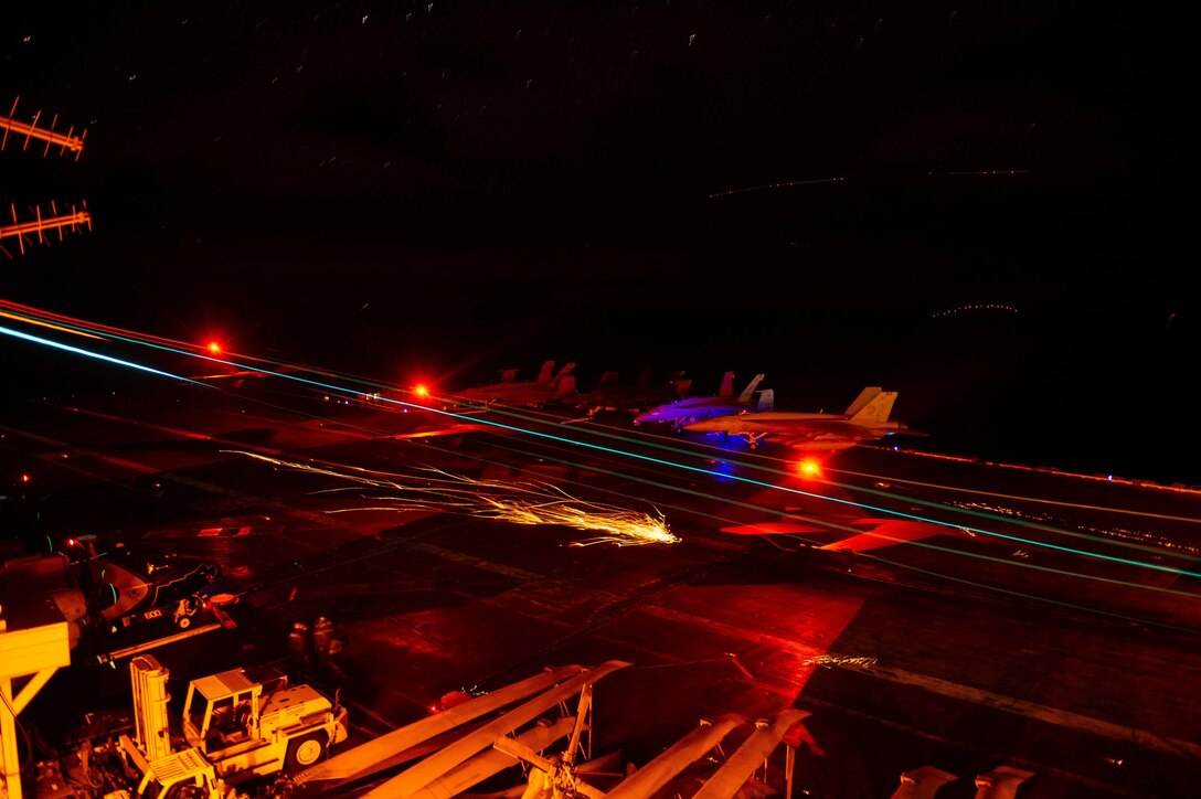 An aircraft sits on the deck of a ship all bathes in red and gold lights.