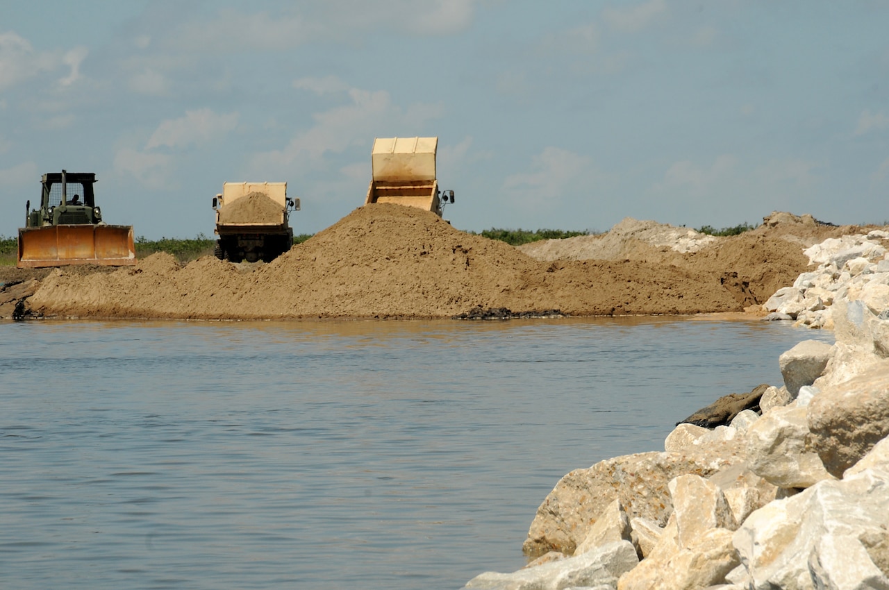 A digger and two dump trucks place soil at the edge of a body of water.