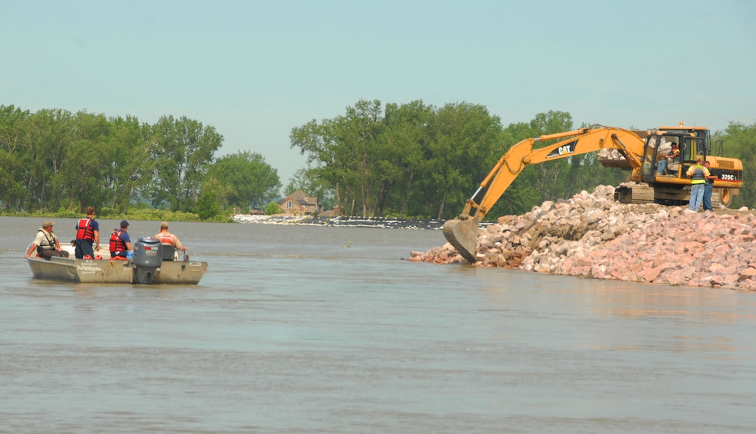 Coast Guardsmen in a small motorboat watch from the water as a backhoe places rocks on a pile.